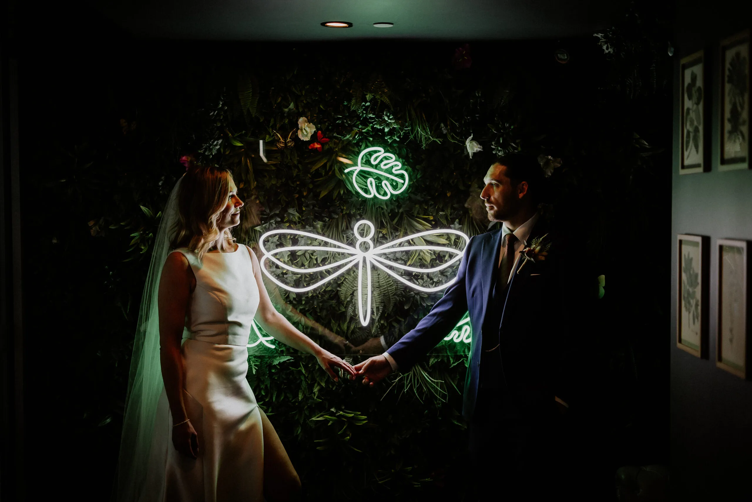Bride and groom holding hands under a neon dragonfly sign