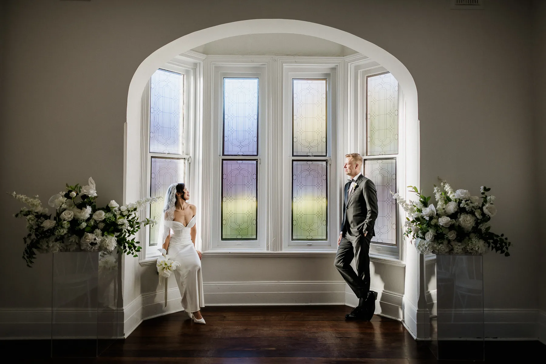 Bride and groom either side of an arched bay window with flowers