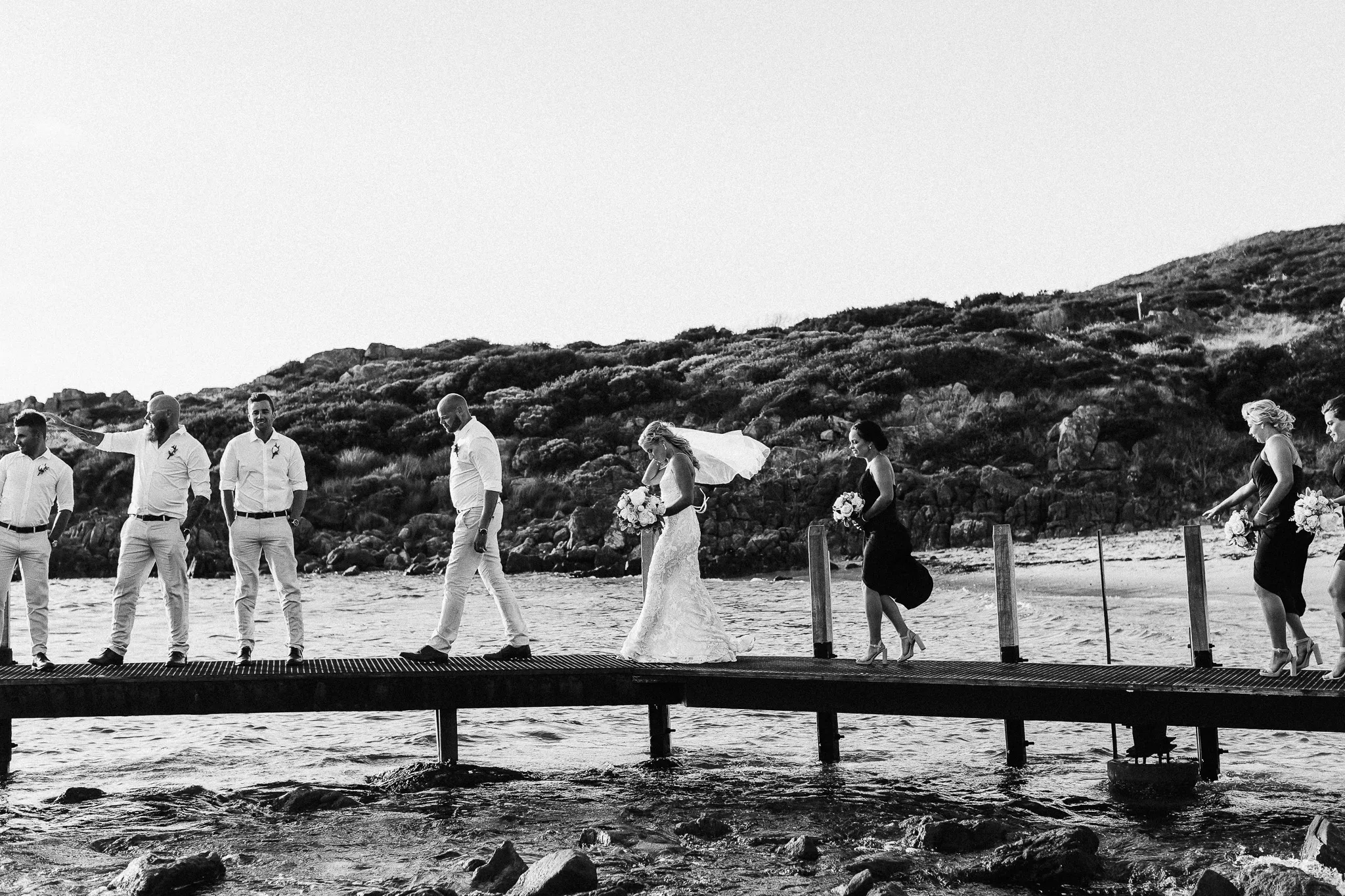 Black and white photo of a wedding party walking along a jetty by the sea