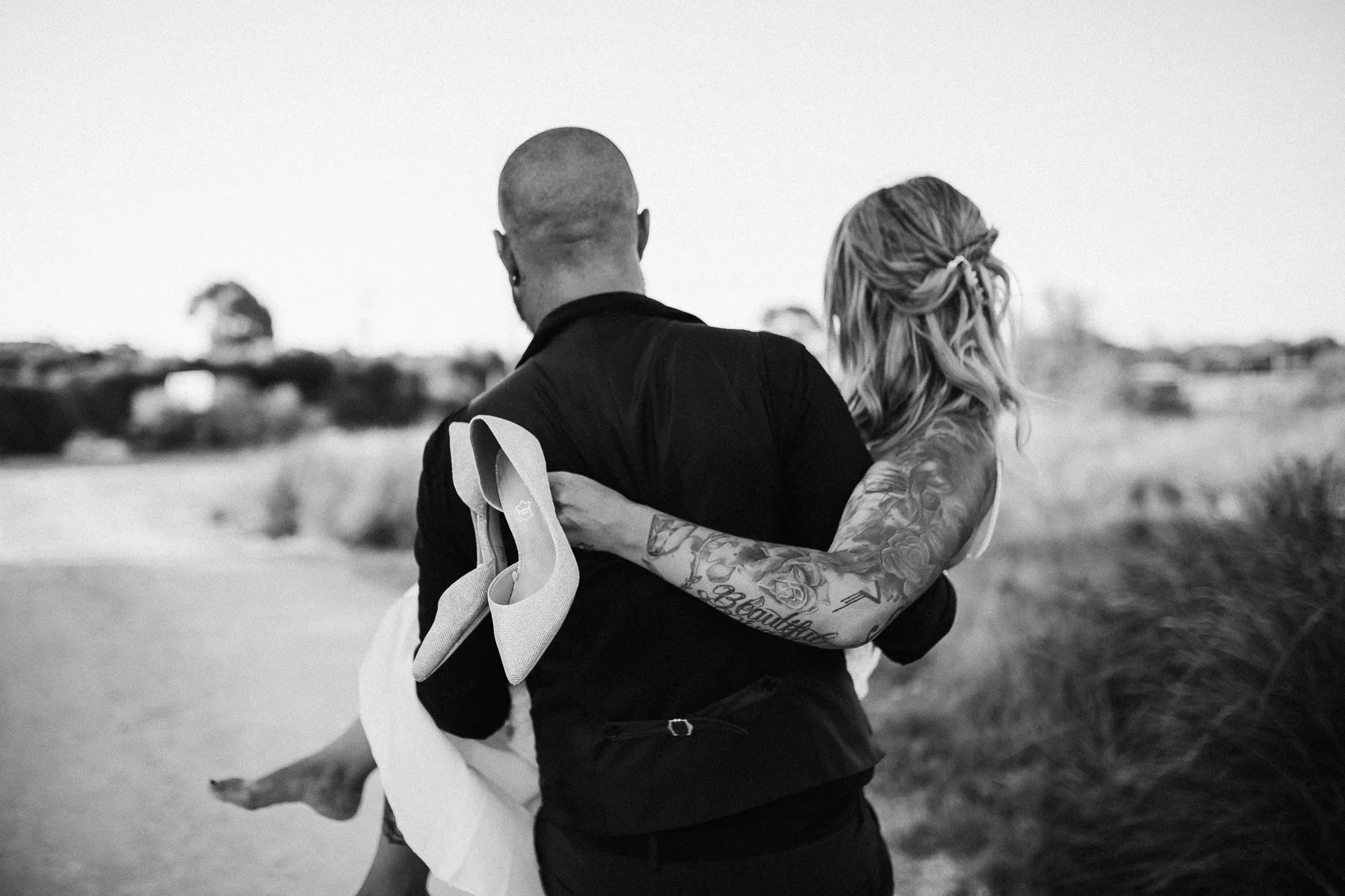 Black and white photo of a groom carrying his barefoot bride, shoes in hand