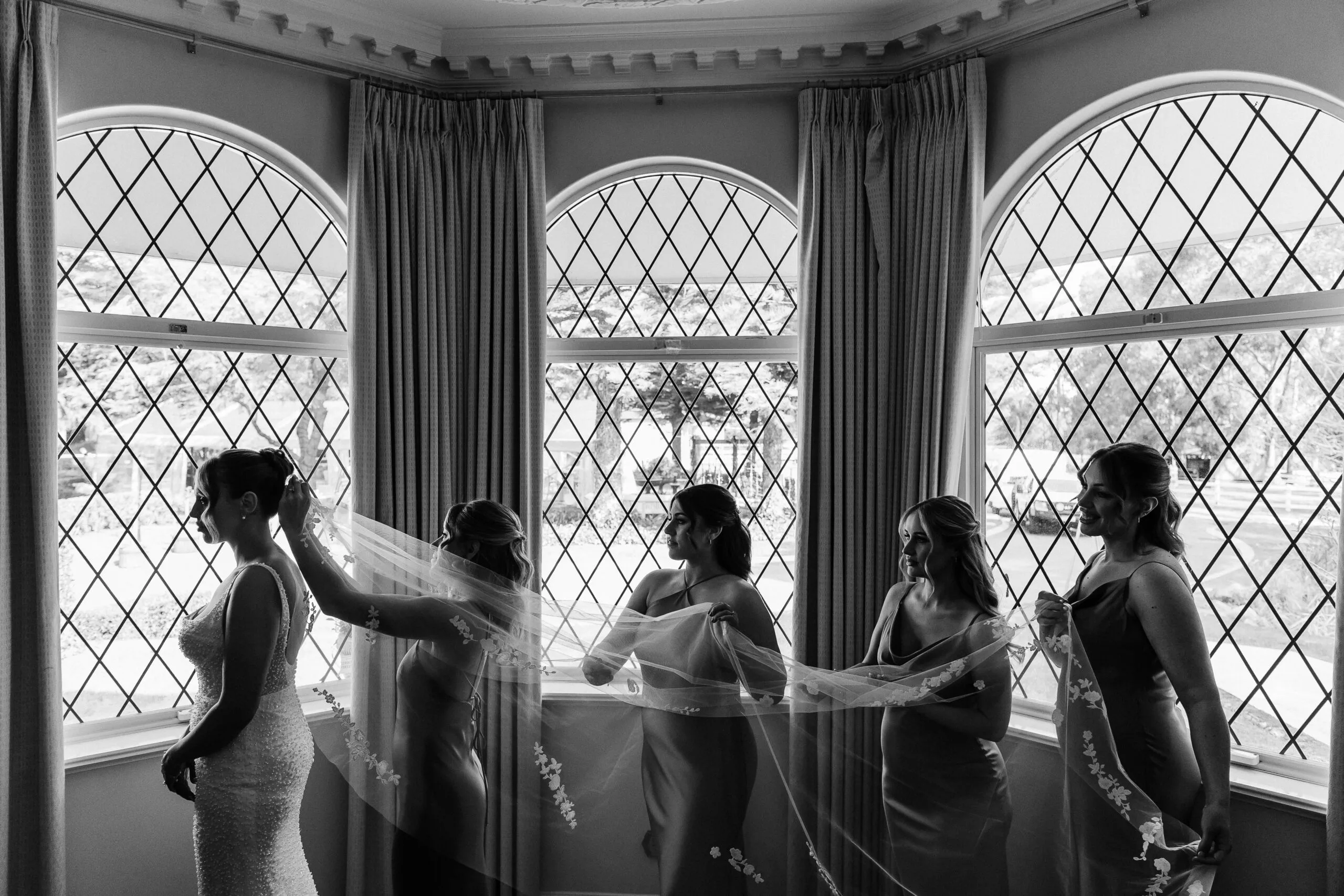 Bridesmaids helping the bride with her veil by tall leadlight windows