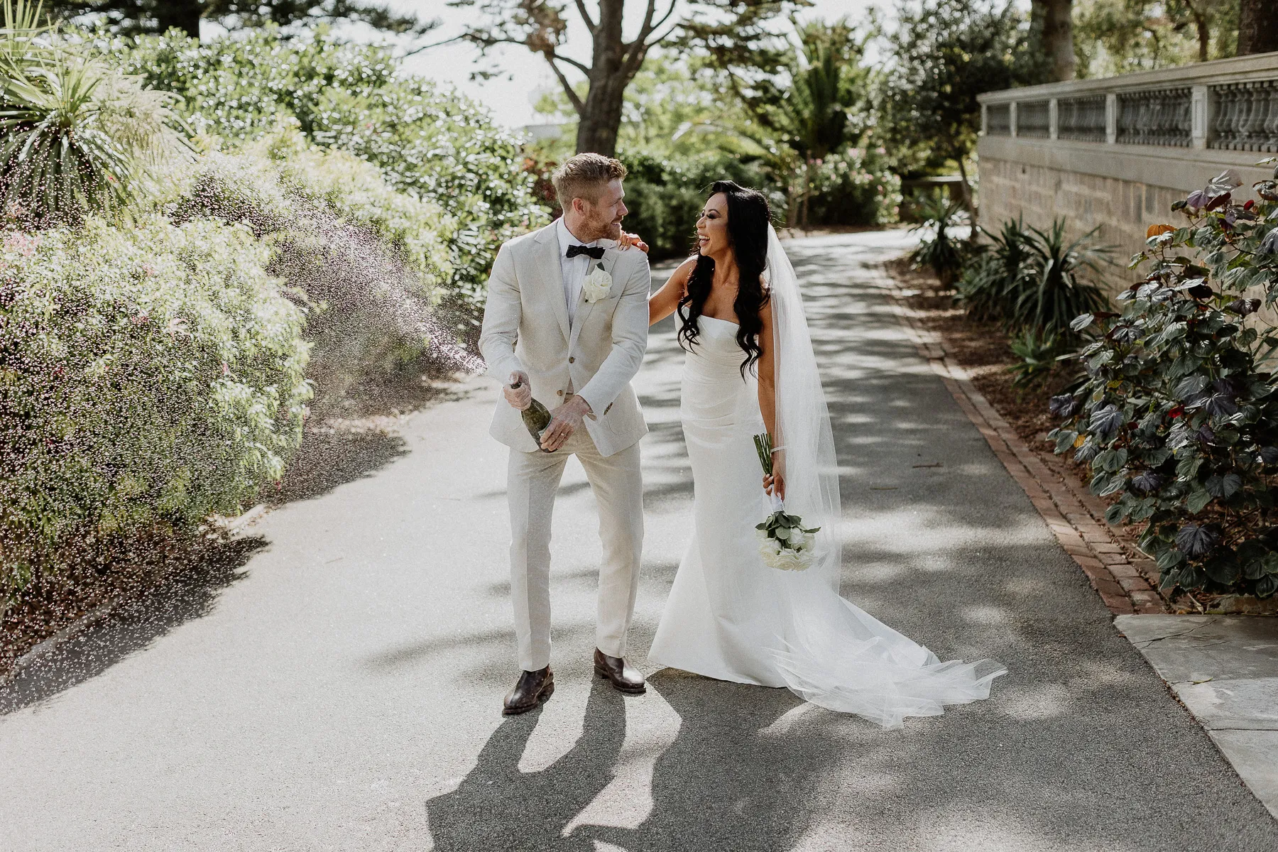 Groom in a white tuxedo and bride on a garden path